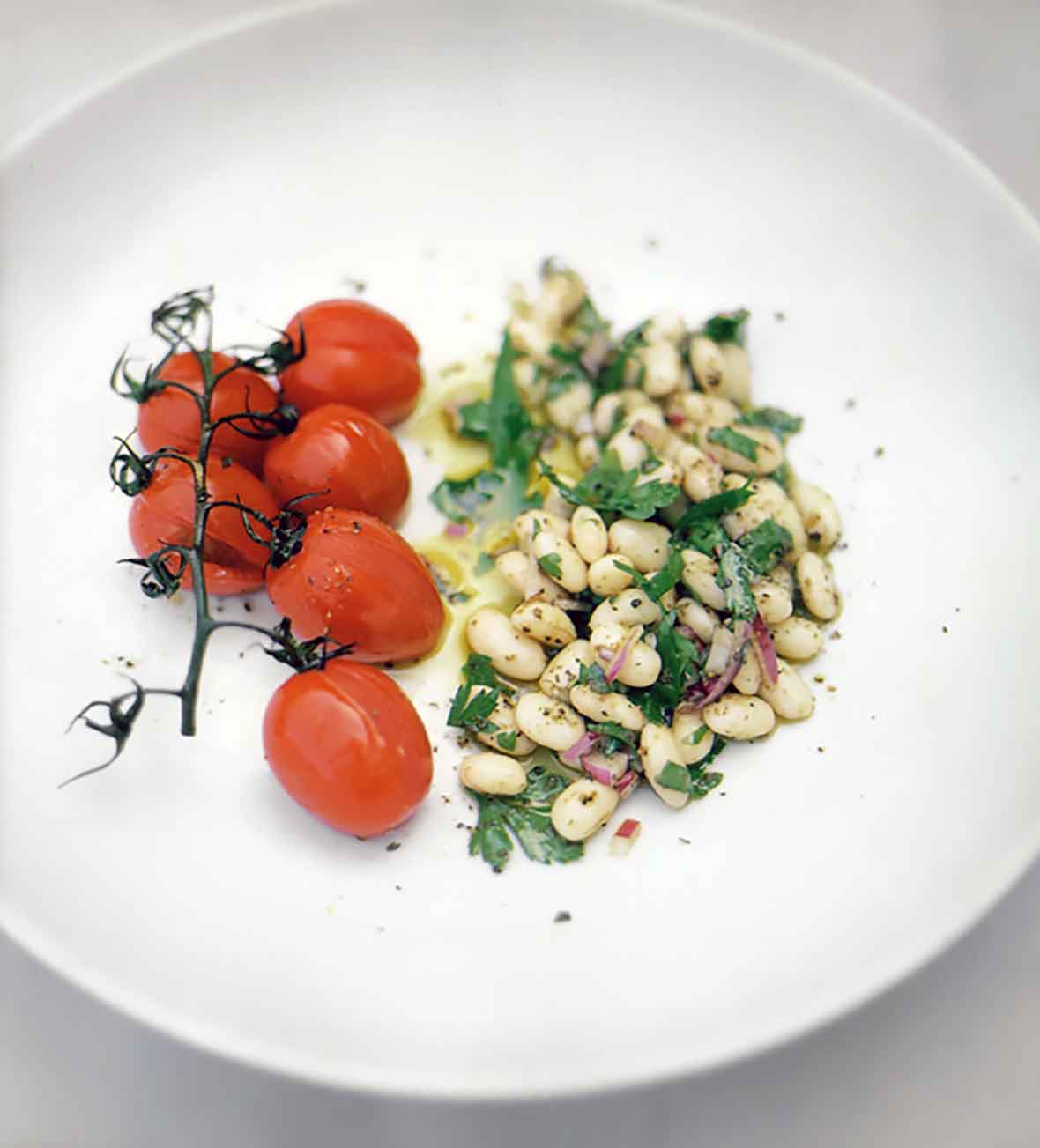 A white plate with a mound of cannellini bean salad and a bunch of grape tomatoes on the vine.