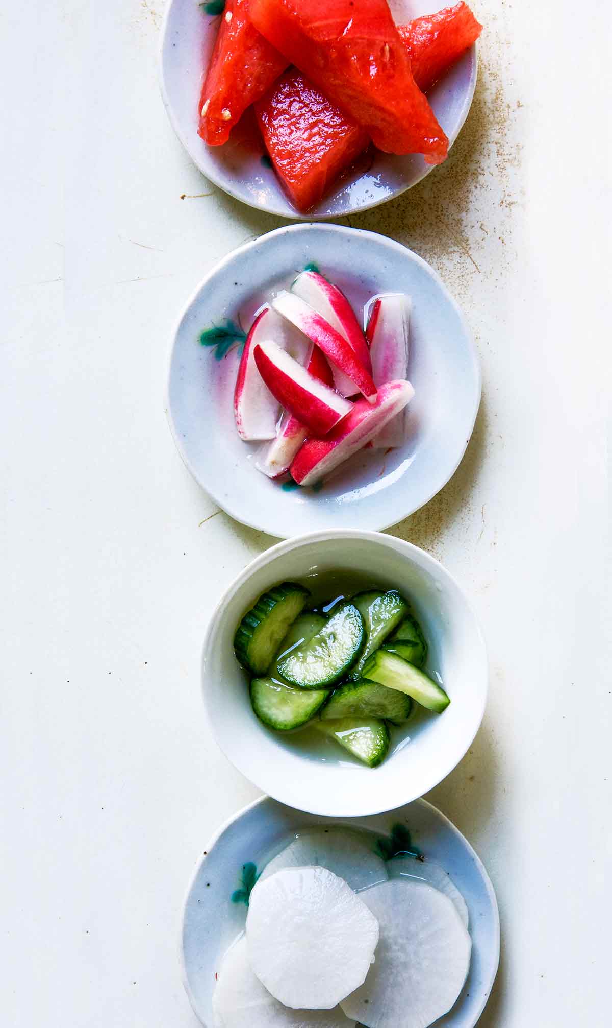 Four bowls of salt and sugar pickles; one with watermelon, one with red radish, one with cucumber, and one with daikon radish.