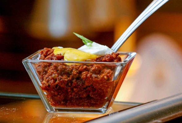 A glass dish of beef chili topped with cheddar cheese sauce and a dollop of sour cream with a spoon resting inside.