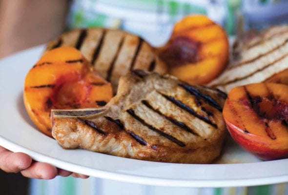A woman holding a white platter of grilled country pork chops with grilled peaches.