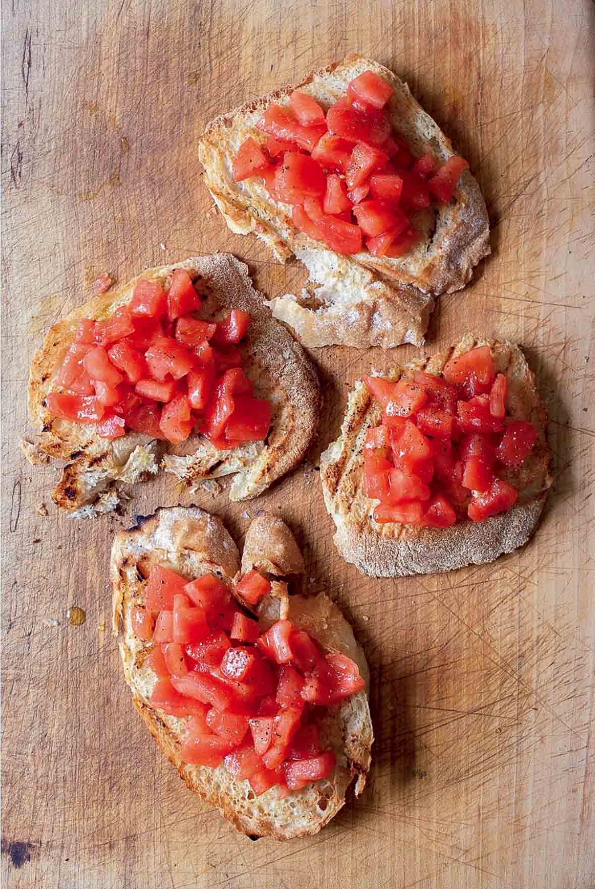 Four slices of bruschetta topped with tomato on a wooden cutting board.