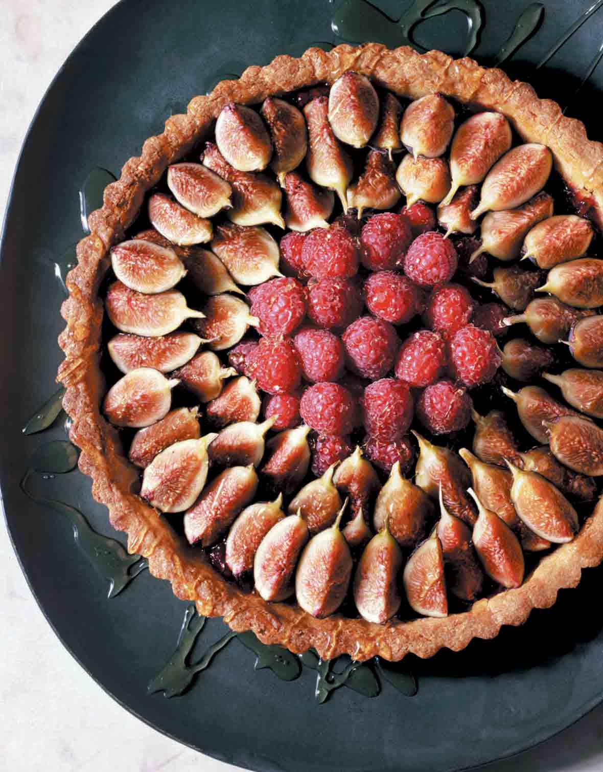 A fresh fig and raspberry tart drizzled with honey on a black platter.