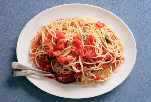 An oval platter topped with a tangle of spaghetti and raw tomatoes with a fork and spoon resting in the pasta.