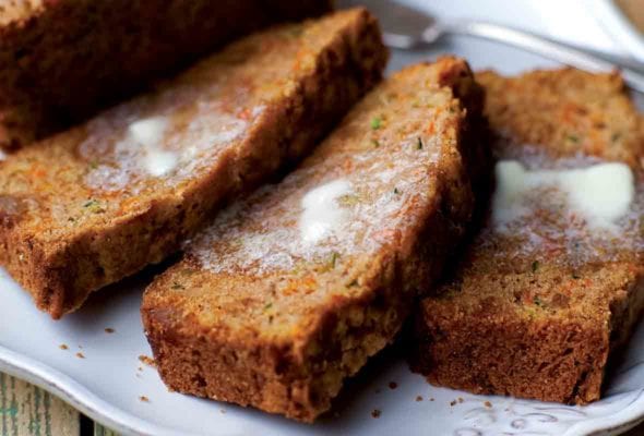 Three buttered slices and an uncut half loaf of carrot zucchini bread with candied ginger on a white platter.