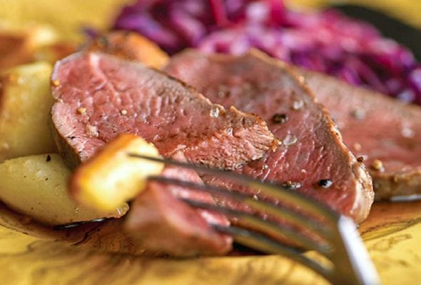Slices of cider-basted venison with roasted potatoes and red cabbage on a yellow plate.