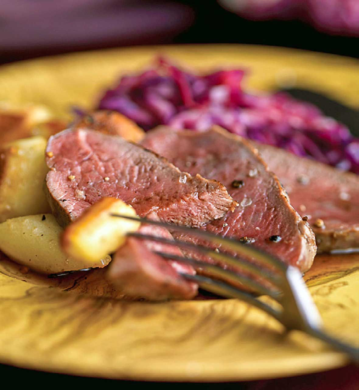 Slices of cider-basted venison with roasted potatoes and red cabbage on a yellow plate.