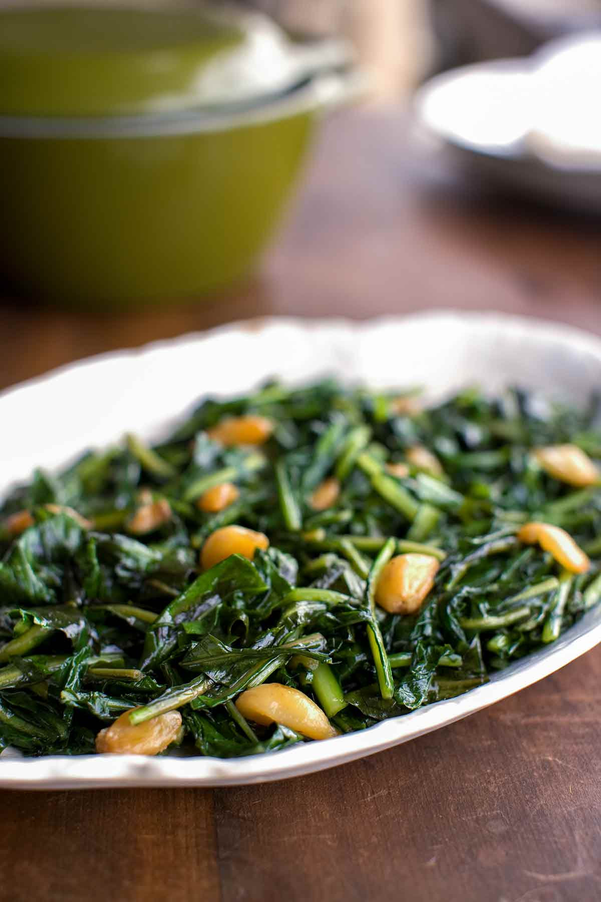 A white oval dish filled with leafy greens and garlic cooked in duck fat on a wooden table.