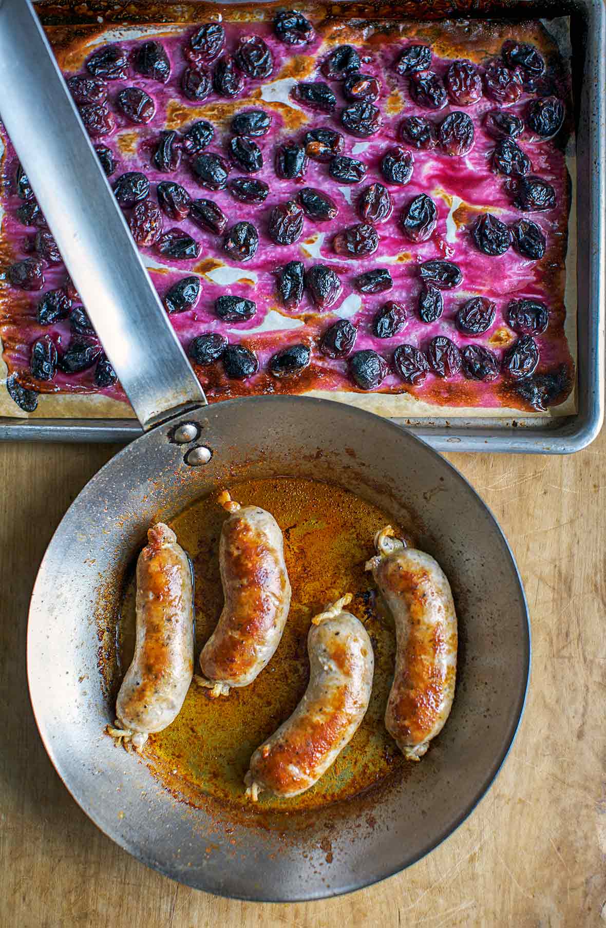Skillet with four seared pork sausages near a baking sheet of roasted grapes