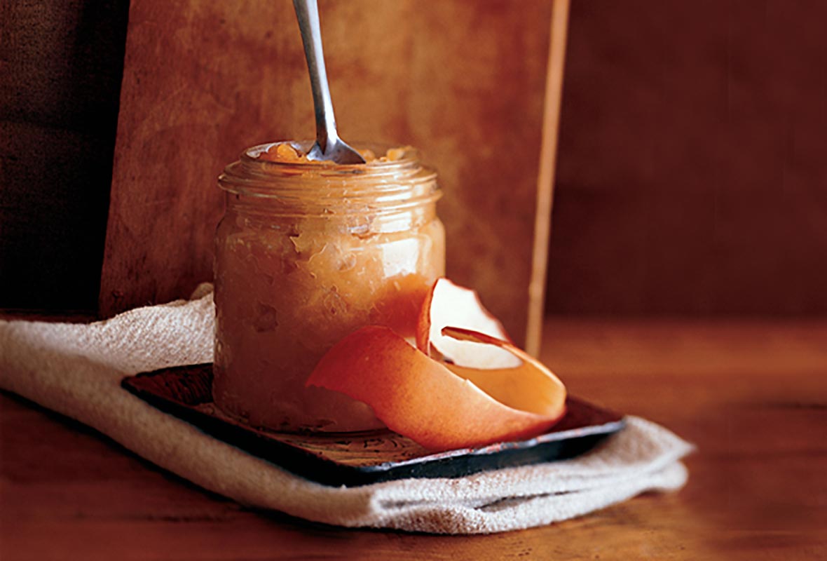 A jar of roasted applesauce with a spoon resting inside on a saucer with a strip of apple peel.
