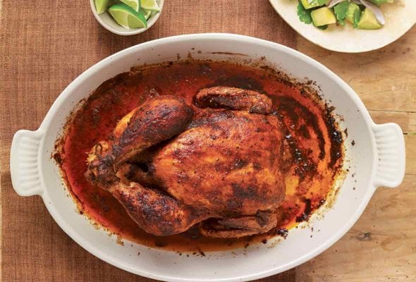 Chicken roasted red with paprika in a casserole dish and plate of lime wedges on a wooden table