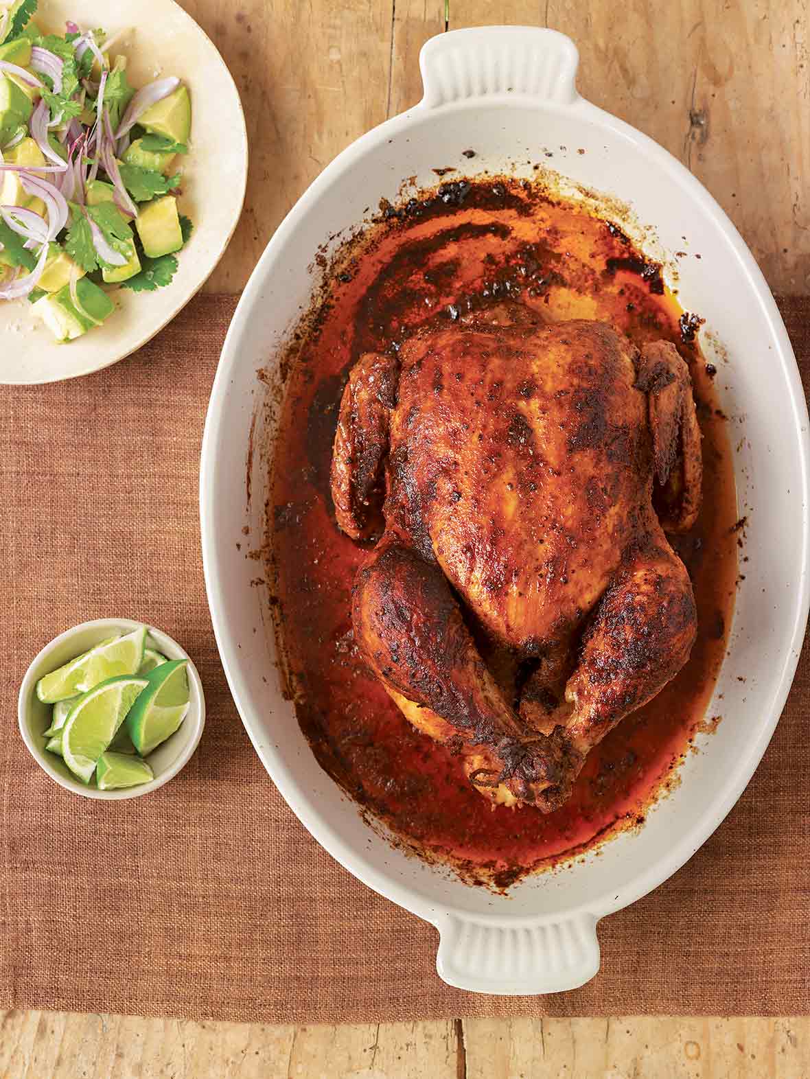 Chicken roasted red with paprika in a casserole dish and plate of lime wedges on a wooden table