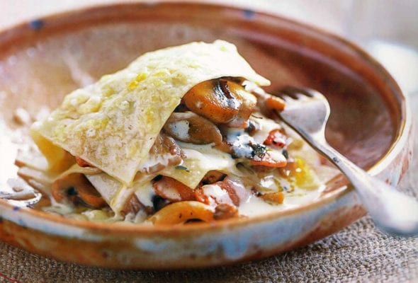 An earthenware bowl with an open lasagna of mushrooms, pine nuts, and thyme in it and a fork resting in the bowl and a glass of white wine in the background.