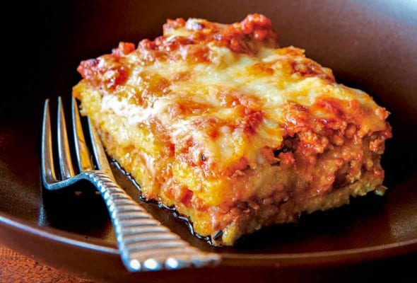 A square of polenta with layers of meat sauce on a plate with a fork, two glasses of wine