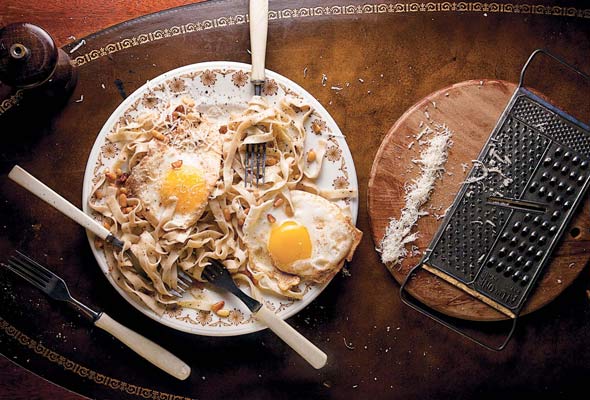 A tangle of brown butter pasta, toasted pine nuts, and two fried eggs on a plate with three forks in it.
