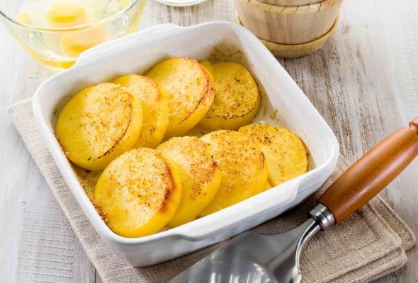 A white casserole dish filled with slices of semolina gnocchi or gnocchi alla Romana on a stack of linen napkins with a serving utensil beside it.