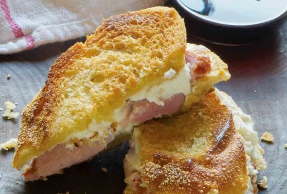 A three-cheese grilled cheese sandwich with ham that is cut in half with a glass of wine in the background.