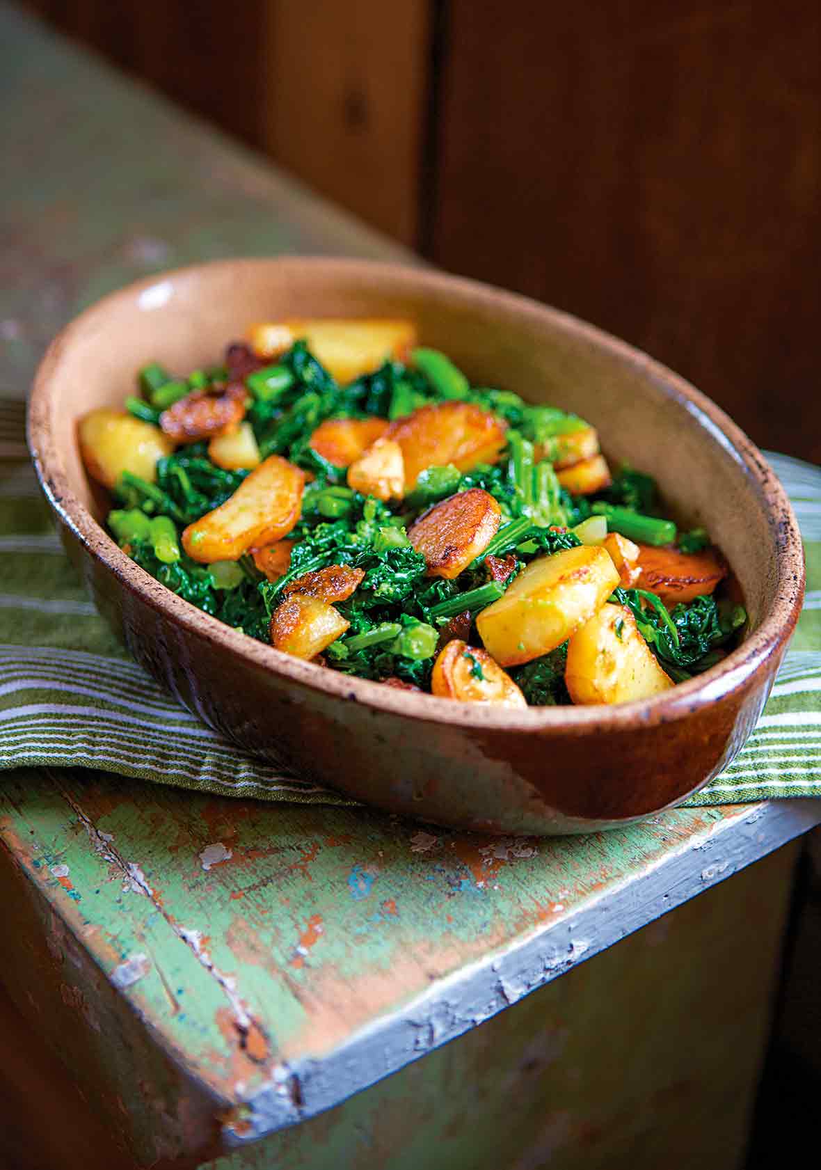 Pieces of sauteed broccoli rabe and slices of seared potatoes in a brown oval dish on a green bench