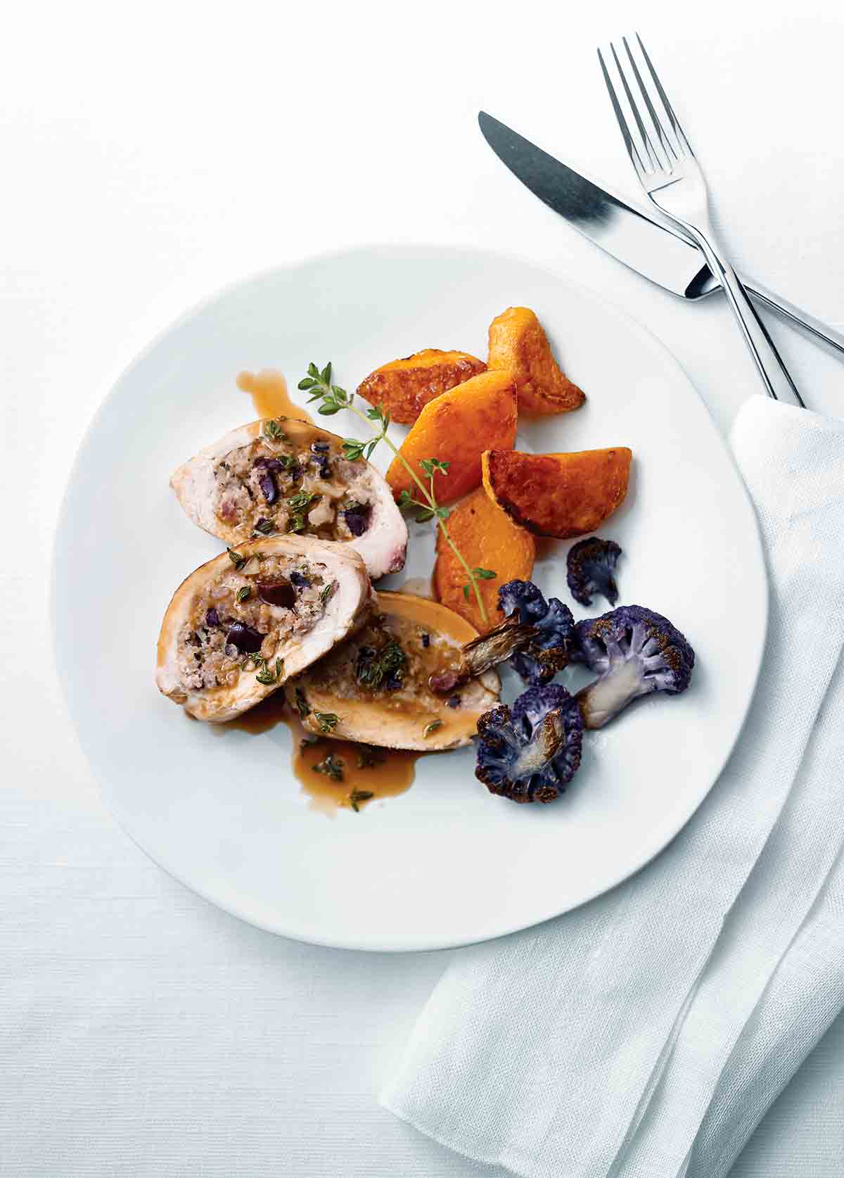 A sliced Mediterranean-style stuffed chicken breast on a white plate with purple cauliflower, sweet potato, and a sprig of thyme.