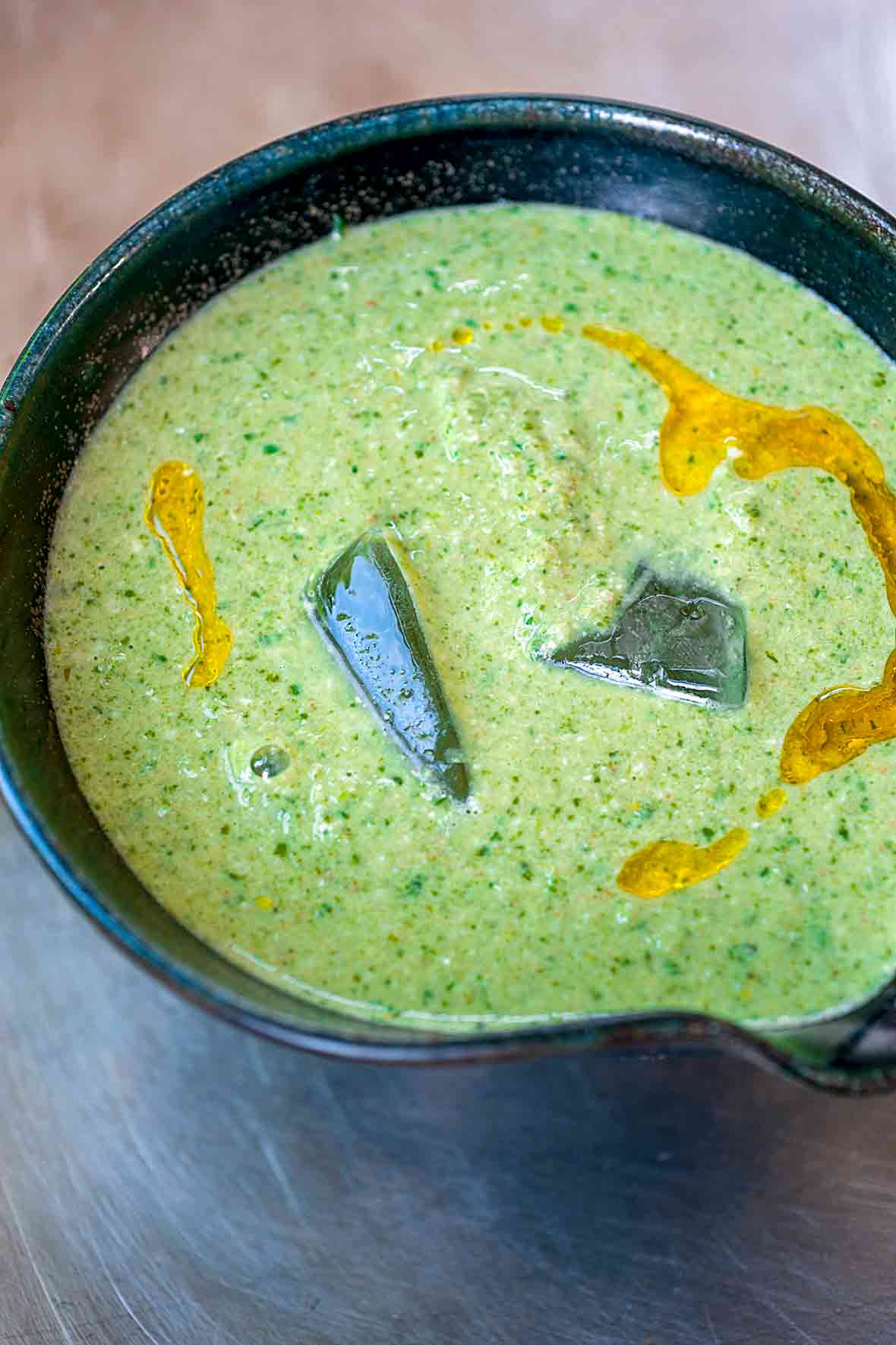Black bowl of green gazpacho, with two ice cubes, drizzle of olive oil