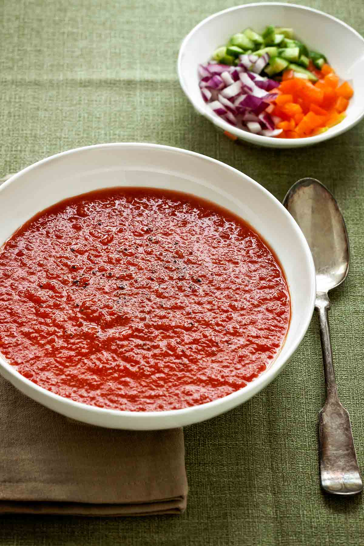 A bowl of Spanish gazpacho on a linen cloth and napkin with a spoon beside it and a bowl of chopped peppers, cucumber, and onion.