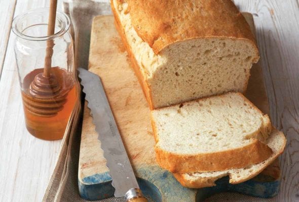 A partially sliced loaf of buttermilk sandwich bread on a wooden cutting board with a bread knife lying beside it.
