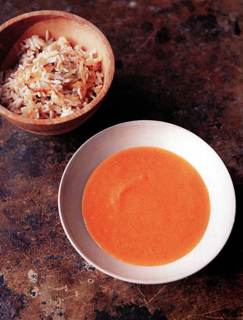 A white bowl of orange carrot and lentil soup with a bowl of rice nearby