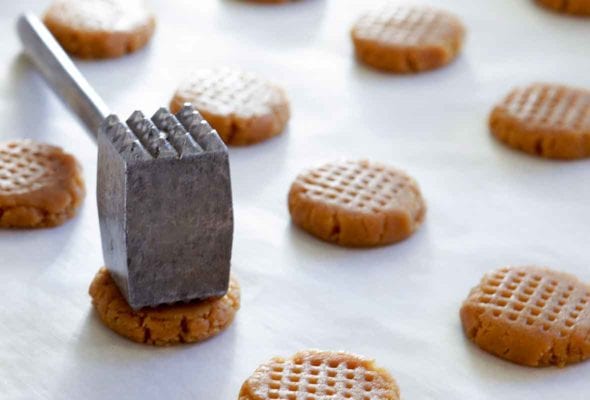 Unbaked crisp peanut butter cookies on a baking sheet being hammered to create cross hatch marks.