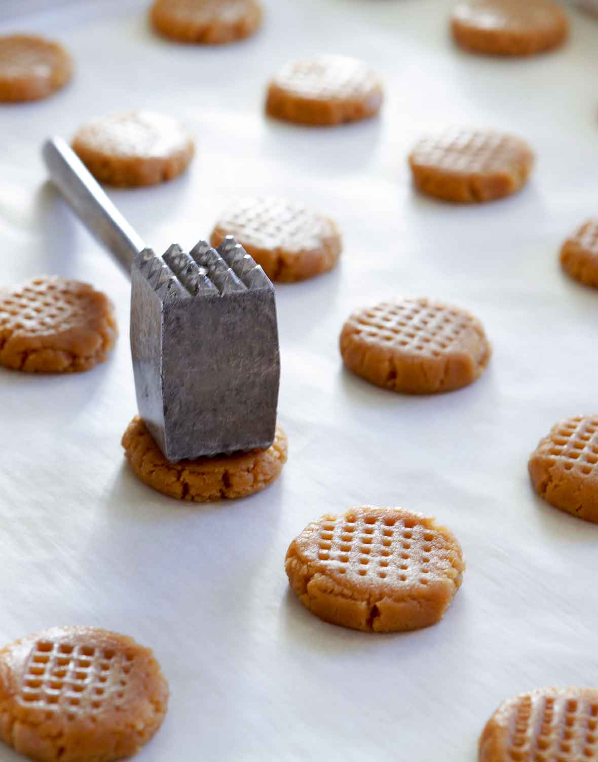 Unbaked crisp peanut butter cookies on a baking sheet being hammered to create cross hatch marks.