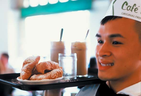 A person carrying a tray with a plate of beignets, two milkshakes, and a glass of water.