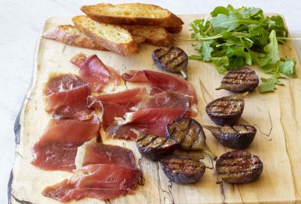 Slices of duck prosciutto, halved grilled figs, arugula, and a few crostini on a wooden serving board.