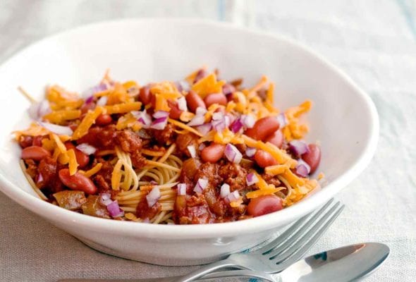 A white bowl of turkey Cincinnati chili with a fork and spoon resting beside it.