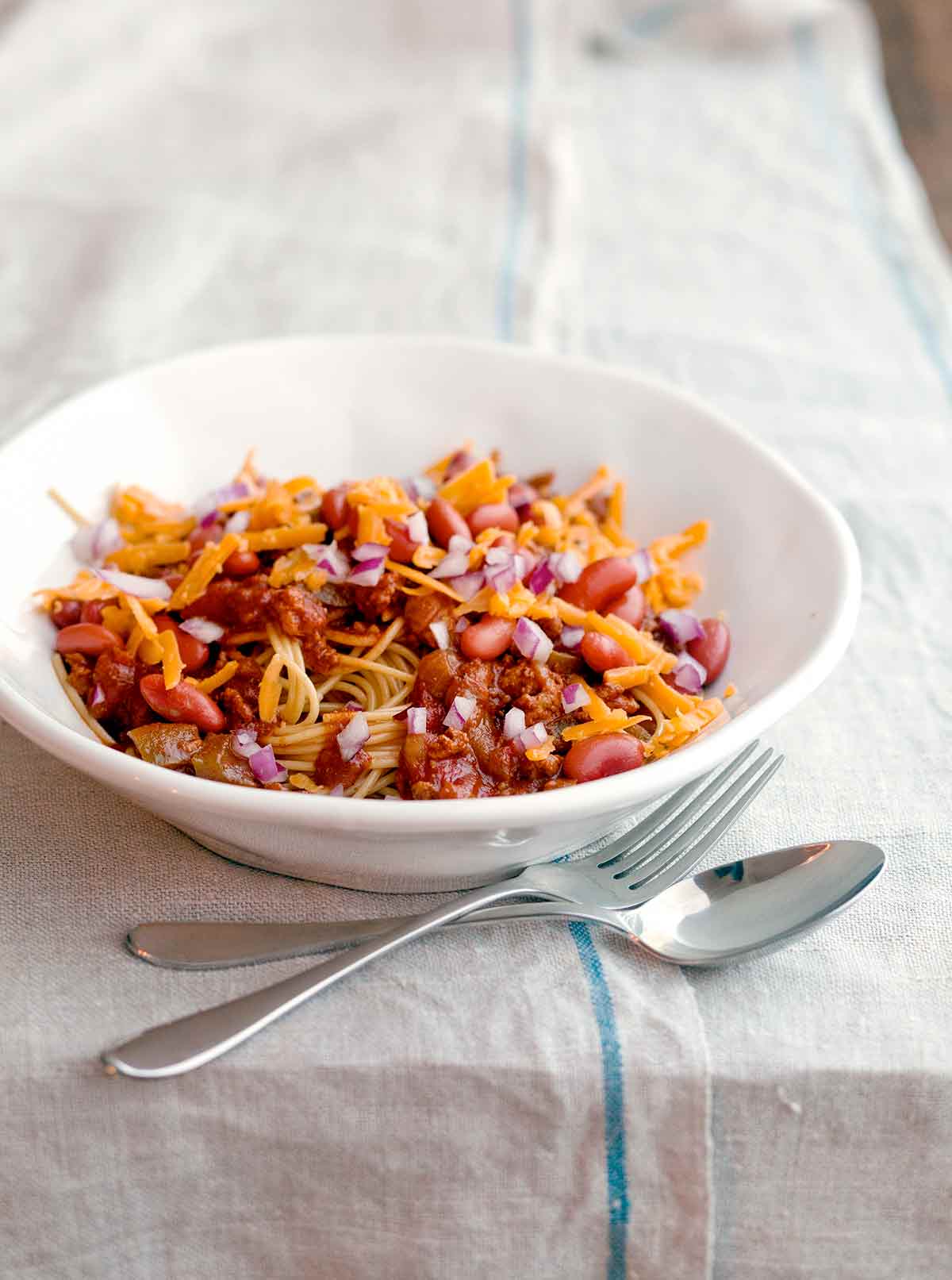 A white bowl of turkey Cincinnati chili with a fork and spoon resting beside it.