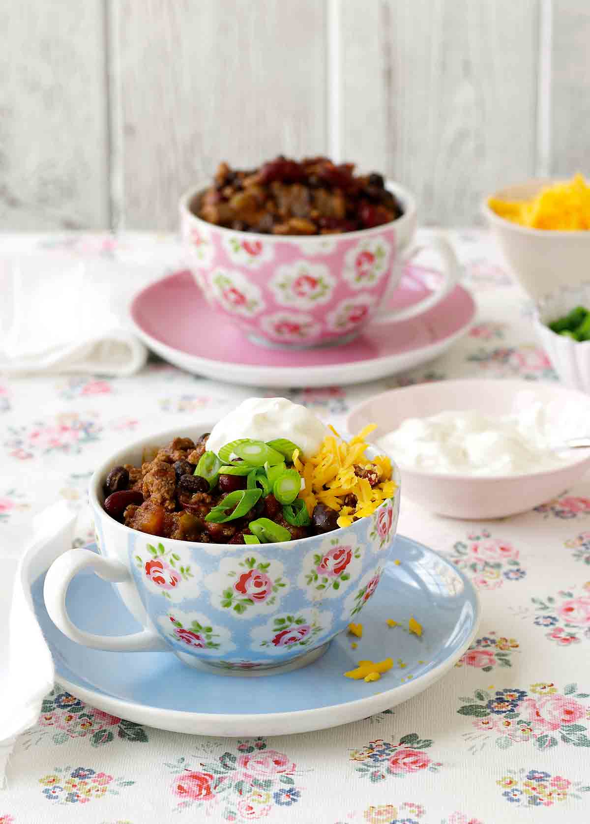 A pretty saucer and teacup filled with game day chili, and topped with scallions, Cheddar, and sour cream.