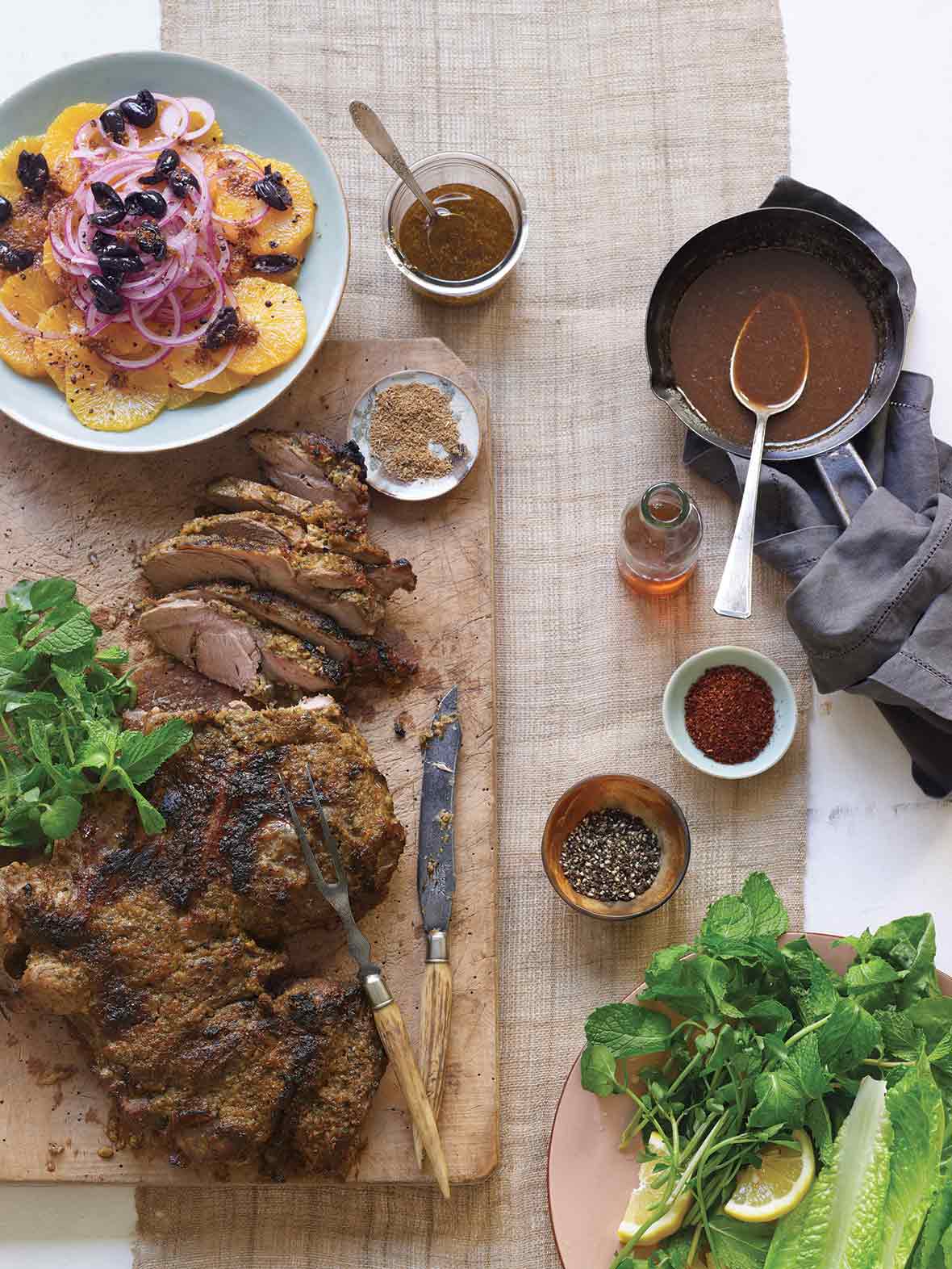A partially carved leg of lamb with Moroccan spices on a wooden board with small dishes of spices, oil, and pan sauce beside it.