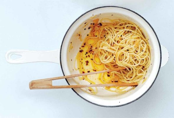 A white enamel pot with a blue rim with spaghetti, chili flakes, and oil in the bottom. A set of wooden tongs sits inside.
