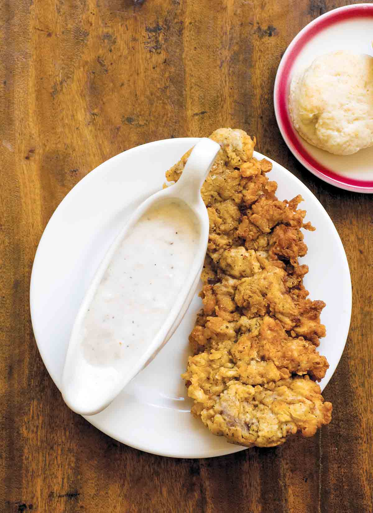 A piece of chicken fried steak with a dish of cream gravy beside it all on a white plate.