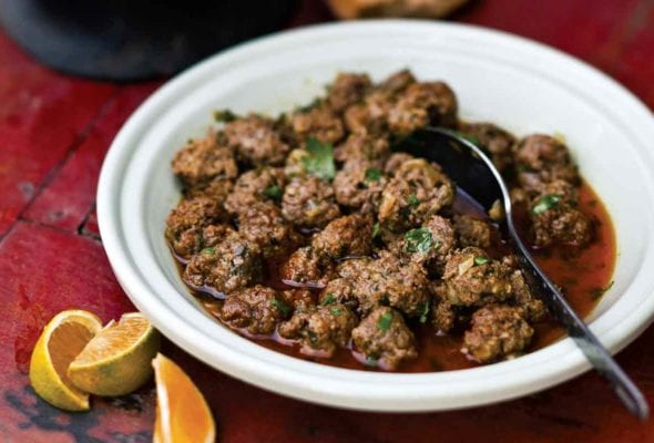 A white bowl filled with Moroccan meatballs with a spoon resting inside and some small orange wedges on the side.