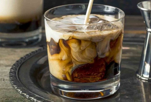 Cream being poured from a pitcher into a highball glass for a white Russian cocktail.