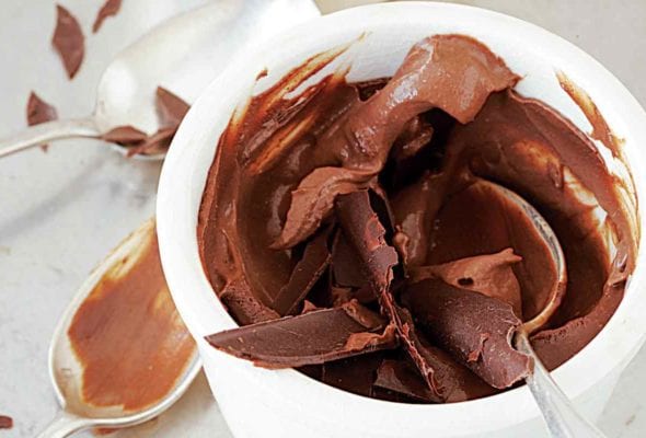 A chocolate pot de crème topped with chocolate shavings in a white cup with two spoons resting beside.