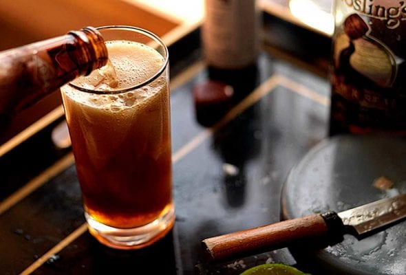 A tall glass filled with dark and stormy cocktail with ginger beer being poured in.