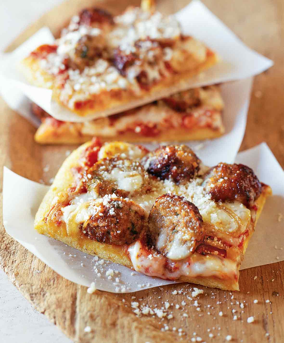 Three square slices of meatball pizza on strips of parchment paper, with fresh Parmesan sprinkled overtop.