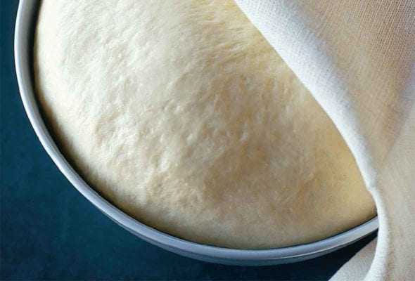 A ball of rising semolina pizza dough in a round metal tin, partially covered by a kitchen towel.
