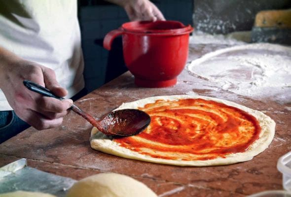 A wooden spoon spreading simple pizza sauce on a round of pizza dough.