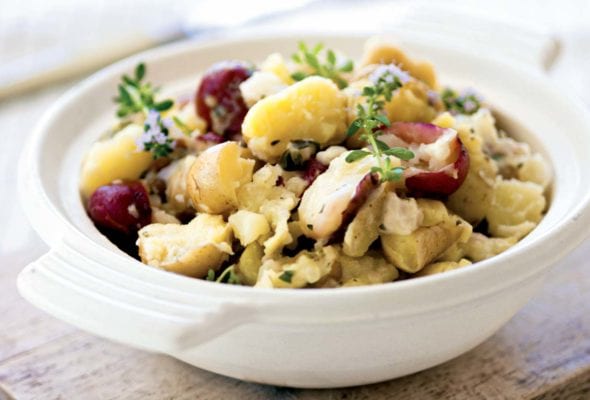 A white serving bowls filled with smashed potatoes and garnished with thyme sprigs on a wooden board.