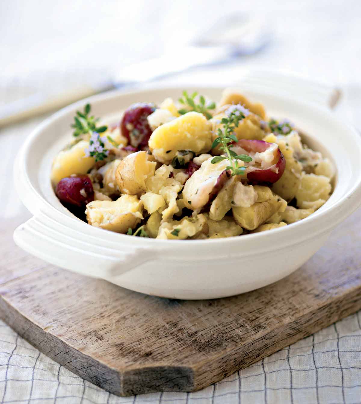 A white serving bowls filled with smashed potatoes and garnished with thyme sprigs on a wooden board.