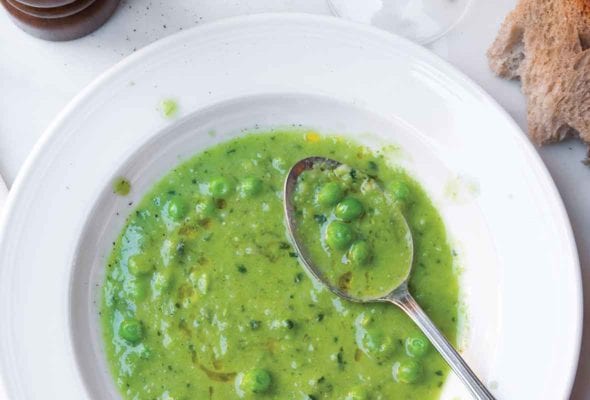 A bowl of pureed pea soup with mint with a spoon resting in the bowl and a glass of wine, piece of bread, and pepper mill beside it.