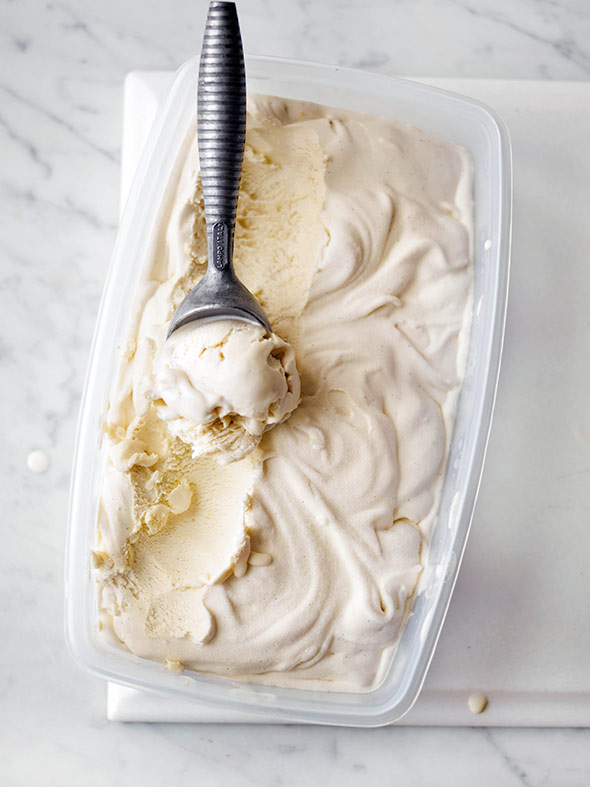 A plastic container filled with vanilla bean ice cream with a metal ice cream scoop resting inside.