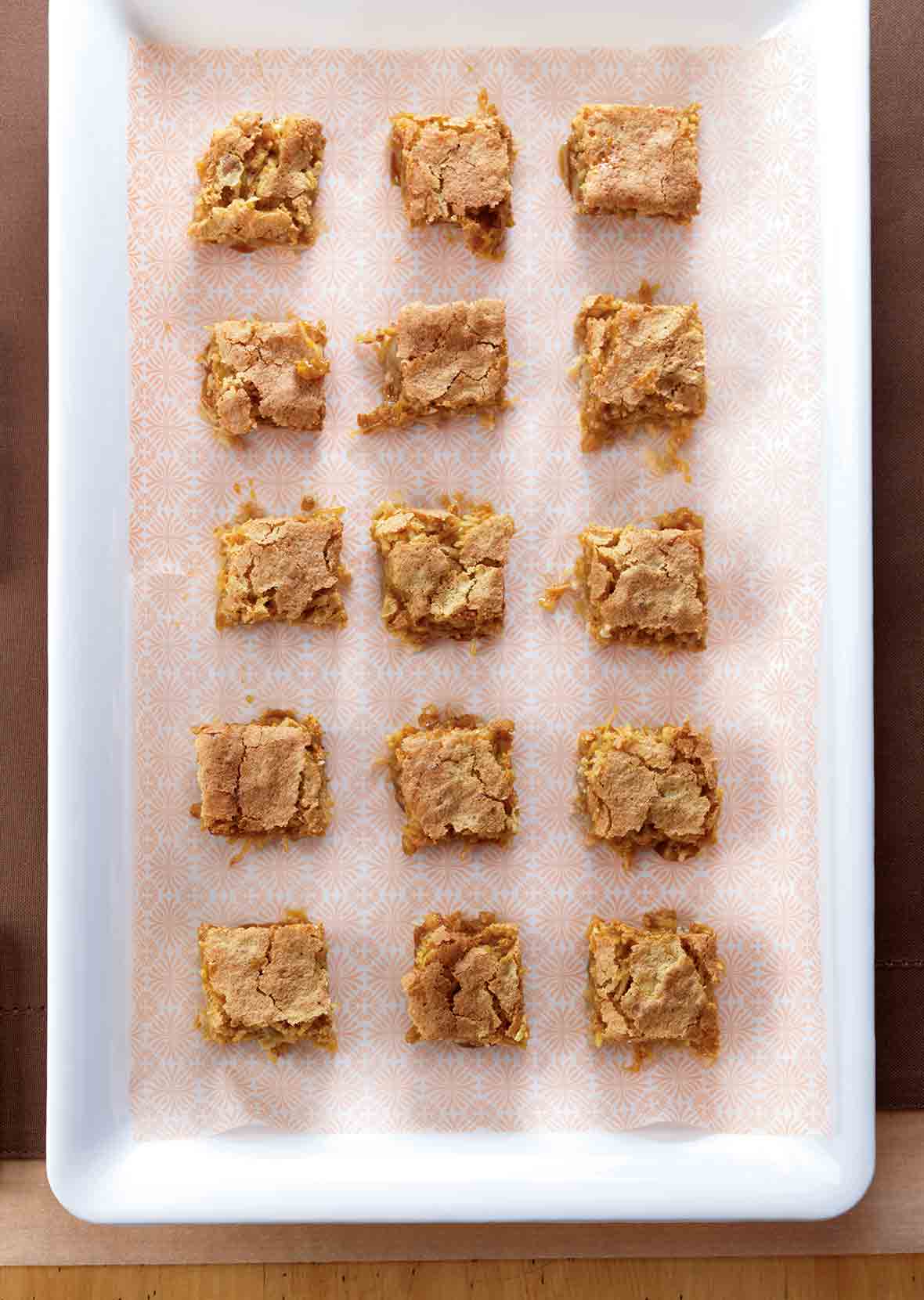 A white baking dish with a piece of patterned parchment and 15 squares of coconut blondies.
