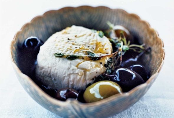 A decorative bowl filled with a sphere of goat cheese and a few olives, topped with a thyme sprig.