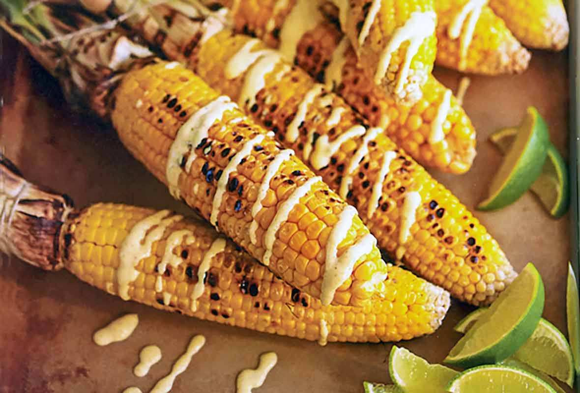 Six ears of grilled Indian corn drizzled with curry yogurt and lime wedges on the side.